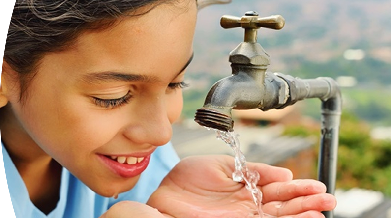 Niña sonriendo mientras recoge agua potable con las manos desde un grifo al aire libre.