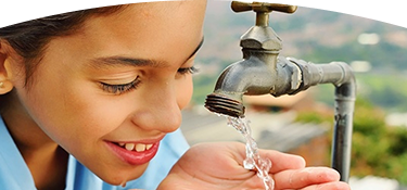 Niña sonriendo mientras recoge agua potable con las manos desde un grifo al aire libre.