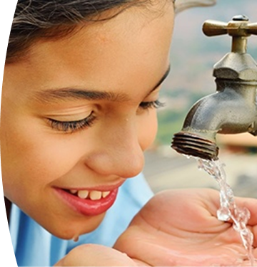 Niña sonriendo mientras recoge agua potable con las manos desde un grifo al aire libre.