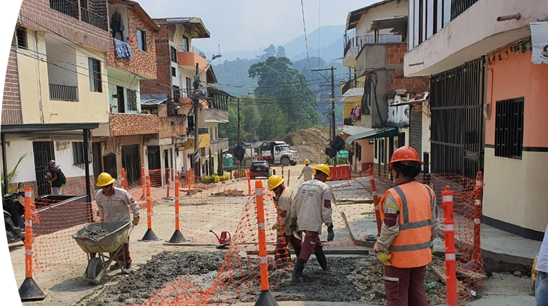 Trabajadores de construcción con cascos y uniformes de seguridad realizan obras en una calle residencial, rodeados de mallas naranjas de protección.