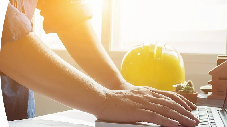 Fotografía de un hombre apoyando sus manos sobre un escritorio mientras trabaja. En el fondo, un casco de construcción amarillo y maquetas de casas. 