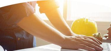 Fotografía de un hombre apoyando sus manos sobre un escritorio mientras trabaja. En el fondo, un casco de construcción amarillo y maquetas de casas. 