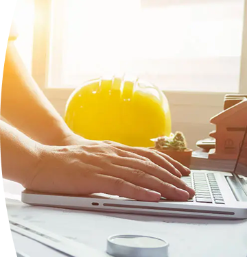 Fotografía de un hombre apoyando sus manos sobre un escritorio mientras trabaja. En el fondo, un casco de construcción amarillo y maquetas de casas. 