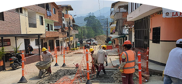 Trabajadores de construcción con cascos y uniformes de seguridad realizan obras en una calle residencial, rodeados de mallas naranjas de protección.