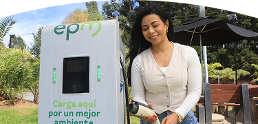 Banner ''Carga pública'' acompañado de una mujer sosteniendo un conector en una estación de carga pública de EPM, rodeada de vegetación