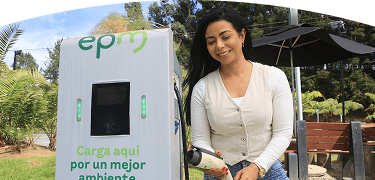 Banner ''Carga pública'' acompañado de una mujer sosteniendo un conector en una estación de carga pública de EPM, rodeada de vegetación