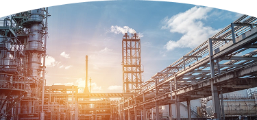 Planta industrial con chimeneas y tuberías, emitiendo vapor o humo bajo un cielo azul con nubes y un sol brillante