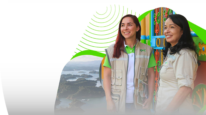 Dos colaboradoras de EPM sonríen durante una visita comunitaria en zona rural, con paisaje montañoso y arquitectura colorida de fondo