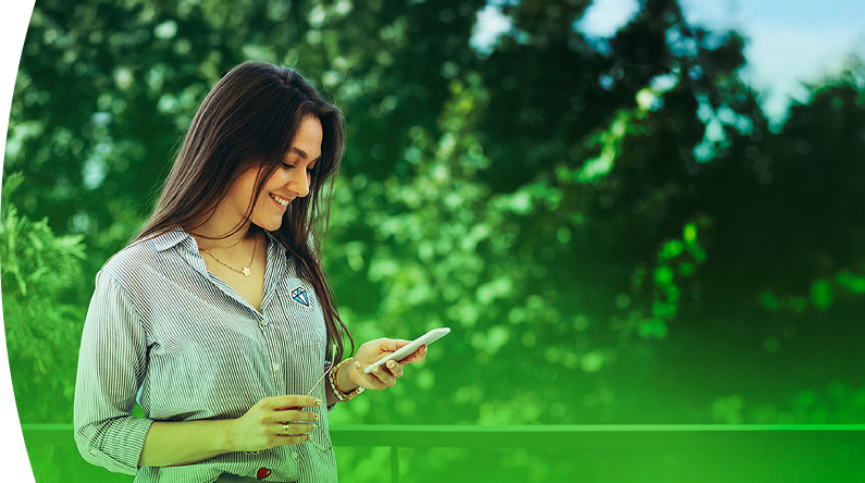 Mujer sonriente con camisa de EPM revisa su celular al aire libre, con fondo verde y naturaleza alrededor