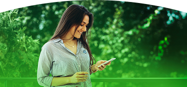 Mujer sonriente con camisa de EPM revisa su celular al aire libre, con fondo verde y naturaleza alrededor