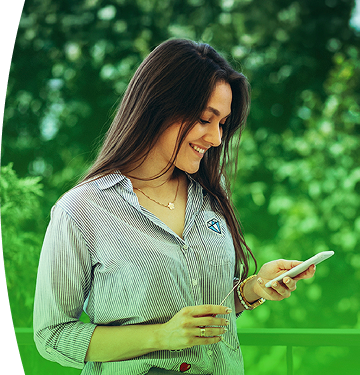 Mujer sonriente con camisa de EPM revisa su celular al aire libre, con fondo verde y naturaleza alrededor