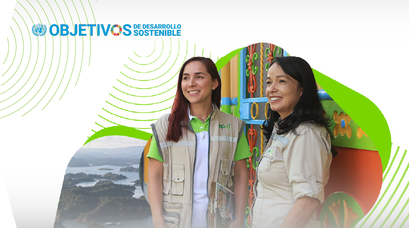 Dos colaboradoras de EPM sonríen durante una visita comunitaria en zona rural, con paisaje montañoso y arquitectura colorida de fondo