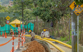 Trabajadores de EPM instalan tuberías amarillas a un costado de la vía en una obra pública, con señalización y medidas de seguridad visibles en el área