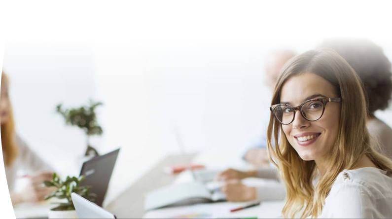 Mujer con gafas y cabello largo sonríe mientras trabaja en una oficina. En el fondo, otras personas están usando computadoras. 