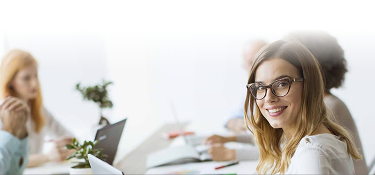 Mujer con gafas y cabello largo sonríe mientras trabaja en una oficina. En el fondo, otras personas están usando computadoras. 