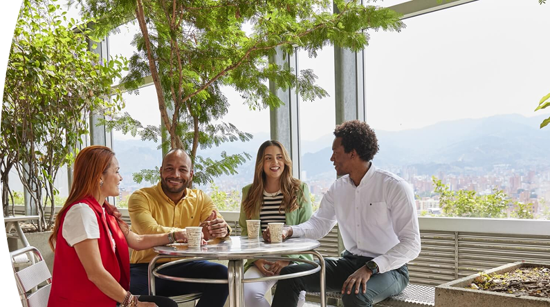 Cuatro personas conversan y disfrutan de un café en terraza con plantas y vista panorámica de la ciudad y montañas