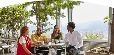 Cuatro personas conversan y disfrutan de un café en terraza con plantas y vista panorámica de la ciudad y montañas