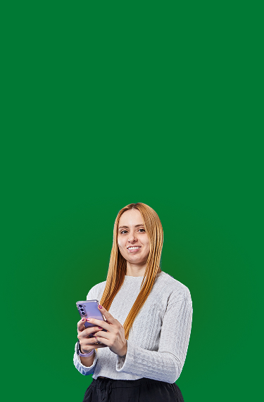 Mujer de cabello largo sostiene un teléfono celular con ambas manos y sonríe a la cámara; viste suéter gris sobre un fondo verde liso