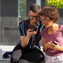 Dos personas jóvenes sonríen mientras miran juntas la pantalla de un celular al aire libre.