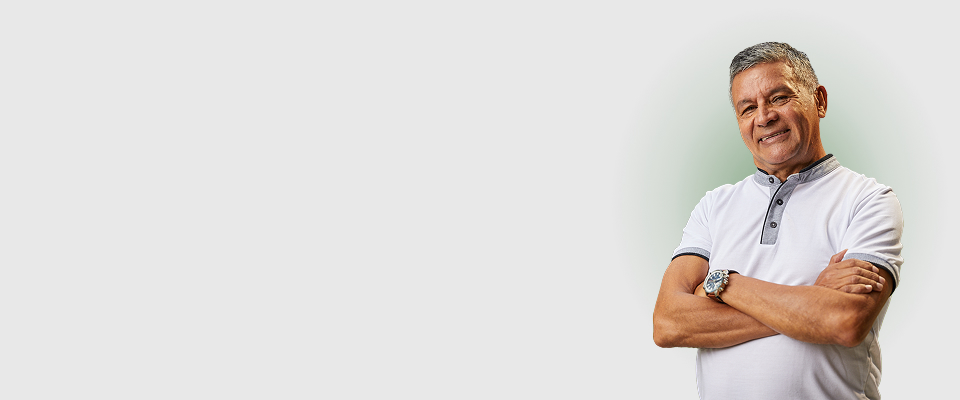 Hombre mayor sonriente con brazos cruzados, vestido con camiseta blanca, posando sobre fondo gris claro