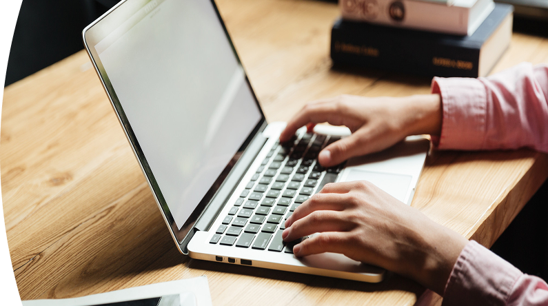 Persona con camisa rosada escribiendo en un computador portátil plateado sobre un escritorio de madera, con libros apilados al fondo