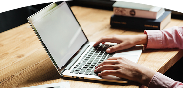Persona con camisa rosada escribiendo en un computador portátil plateado sobre un escritorio de madera, con libros apilados al fondo