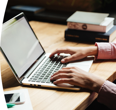 Persona con camisa rosada escribiendo en un computador portátil plateado sobre un escritorio de madera, con libros apilados al fondo