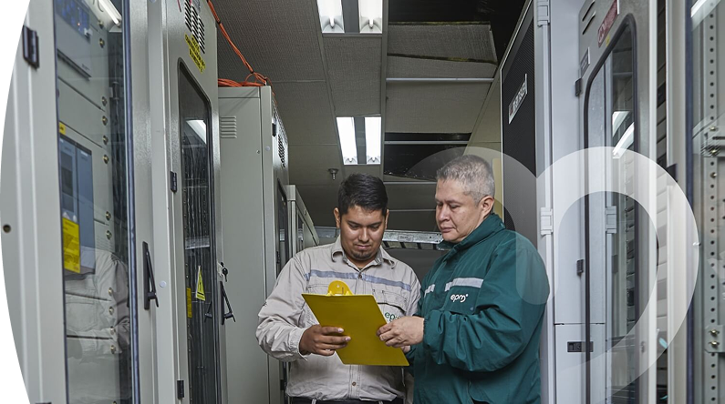 Dos personas en un entorno industrial, una revisando un portapapeles amarillo con equipos técnicos en el fondo.