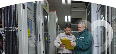 Dos personas en un entorno industrial, una revisando un portapapeles amarillo con equipos técnicos en el fondo.