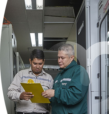 Dos personas en un entorno industrial, una revisando un portapapeles amarillo con equipos técnicos en el fondo.