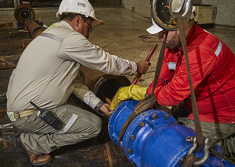 Dos trabajadores con casco y uniforme, uno beige y otro rojo, realizan labores de ajuste en una tubería azul de gran tamaño