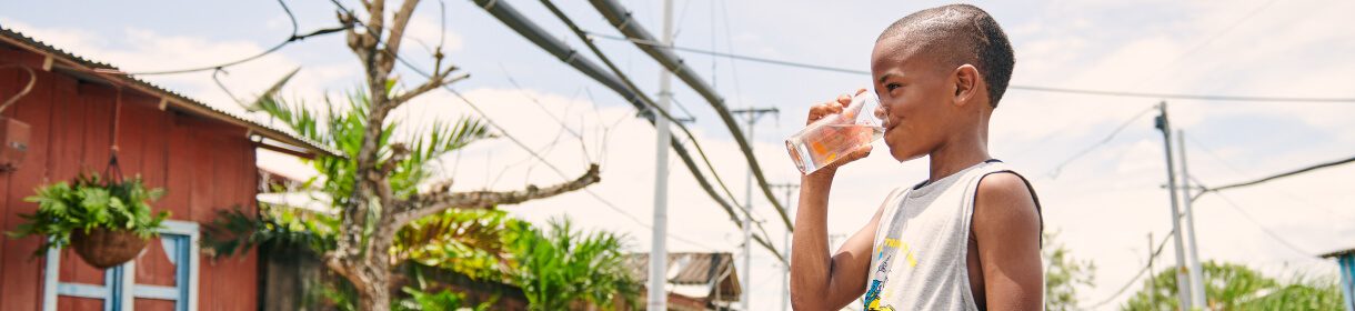 Un niño sonriente bebe un vaso de agua potable frente a una vivienda de madera en un entorno rural iluminado por el sol