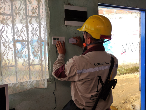 Técnico con casco y uniforme de contratista instala o revisa un medidor eléctrico en el interior de una vivienda.