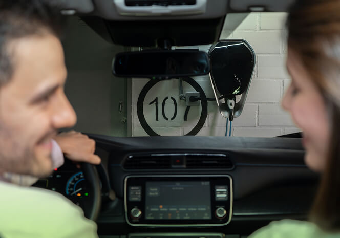 Pareja dentro de un vehículo eléctrico se observa sonriente mientras el carro se carga con el punto de carga número 107 al fondo.
