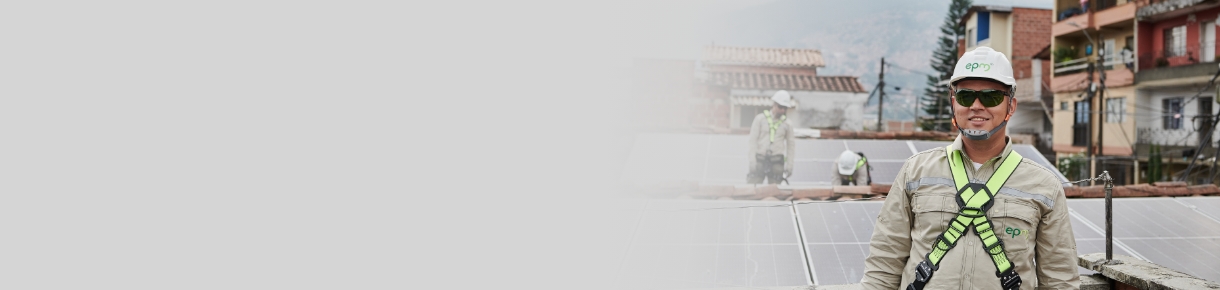 Trabajador con casco y arnés de seguridad realizando instalación de paneles solares en un techo, con más técnicos al fondo en un entorno urbano