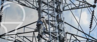 Un técnico realizando mantenimiento en un torre de transmisión eléctrica, escalando por una estructura metálica con cables eléctricos visibles en el fondo