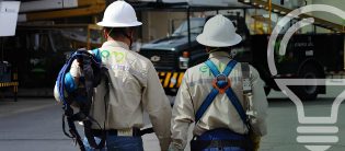 Dos operarios de EPM con cascos y arneses caminan frente a camiones de la empresa en una zona industrial.