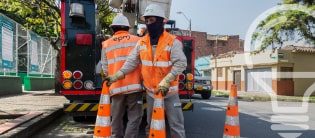Dos trabajadores de la construcción en uniformes naranjas y cascos, de pie cerca de señales de advertencia y una camioneta, en una calle de la ciudad.