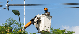 Trabajador de servicios públicos realizando el mantenimiento de un poste de alumbrado público desde una grúa canasta, bajo un cielo despejado