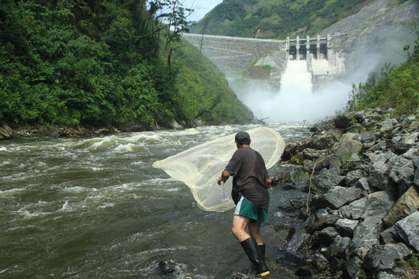 Hombre pescando con atarraya en un río caudaloso, con una represa hidroeléctrica liberando agua al fondo