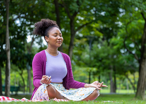 Mujer sentada en posición de meditación sobre el pasto en un parque rodeado de árboles