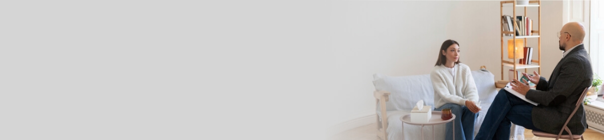 Psicólogo atiende a una paciente en consulta privada, sentados en una sala acogedora con sofá y estantería