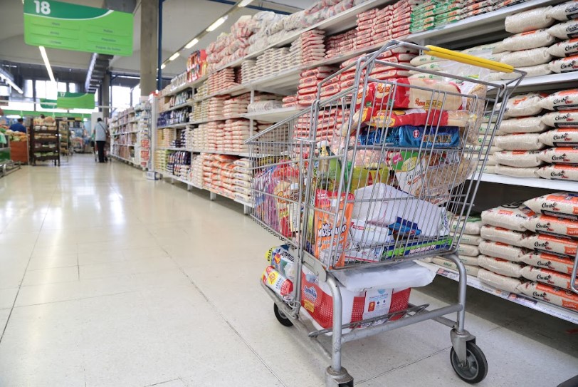 Imagen de un carrito de compras lleno de productos en el pasillo de un supermercado con estantes de arroz y productos básicos.