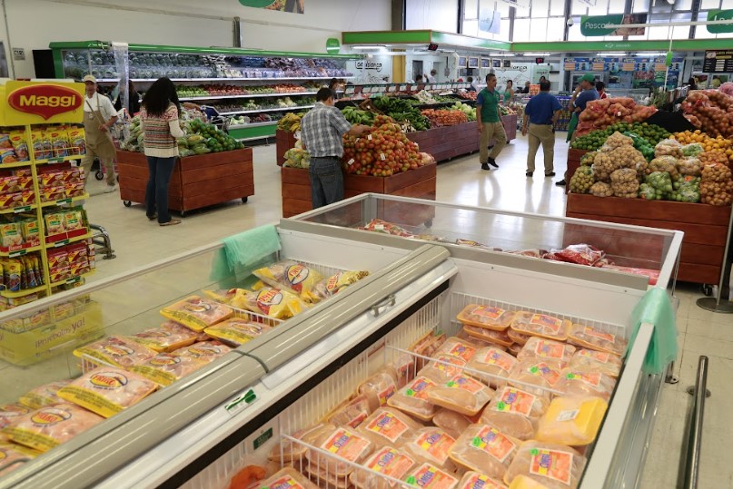 imágen de la vista del área de carnes y productos refrigerados en un supermercado, con estantes llenos de productos y compradores en el fondo.