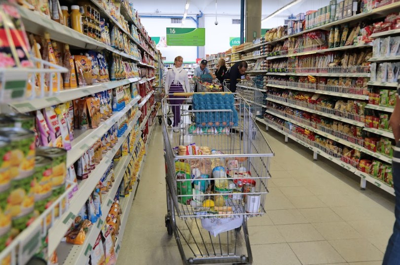 Pasillo del supermercado con un carrito de compras lleno de productos, rodeado de estanterías con artículos enlatados y empacados