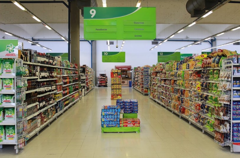 Pasillo de supermercado con estanterías de vinos y mecatos organizadas y un cartel verde con información colgando del techo