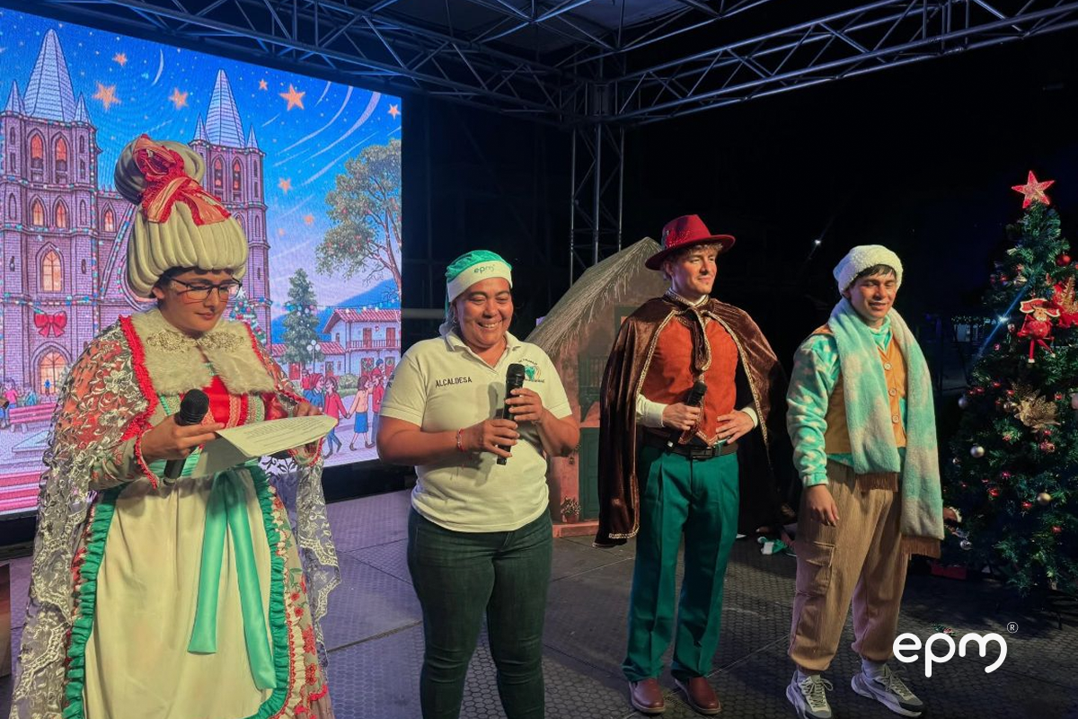 Personas vestidas con ropa navideña interpretando obra de teatro. De fondo se observa pantalla con imágenes de navidad
