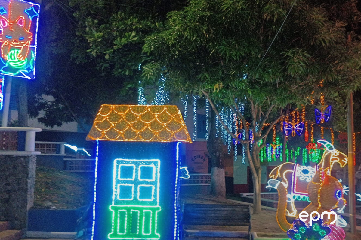 Figura de casa navideña de color amarillo, azul y verde alumbrando rodeada de árboles y figuras navideñas encendidas