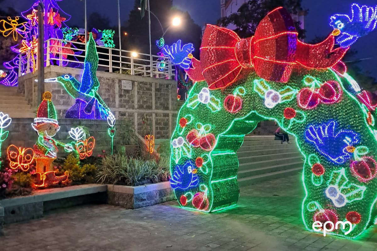 Alumbrando de arco navideño con moño de color rojo en la parte superior. De fondo se observan figuras navideñas alumbrando