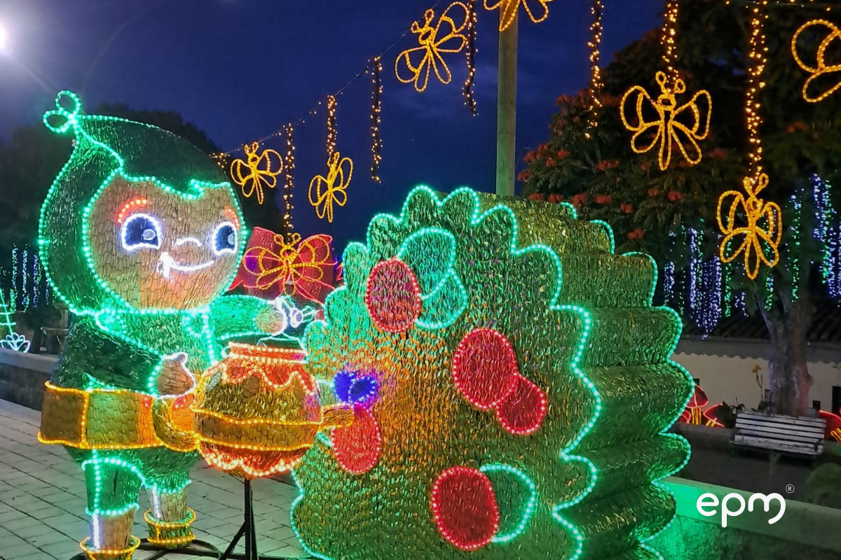 Fotografía de alumbrado navideño en color verde con detalles rojos. De fondo se observan luces amarillas